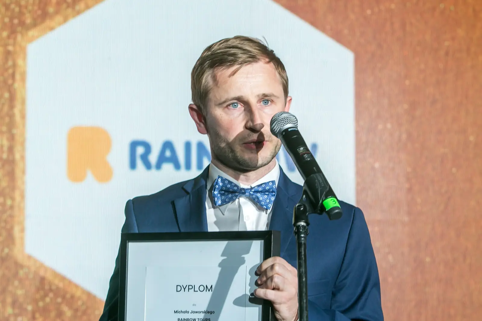 Laureat w niebieskiej muszce w kropki przemawia przy mikrofonie, trzymając dyplom — Gala DMR XI (2022)