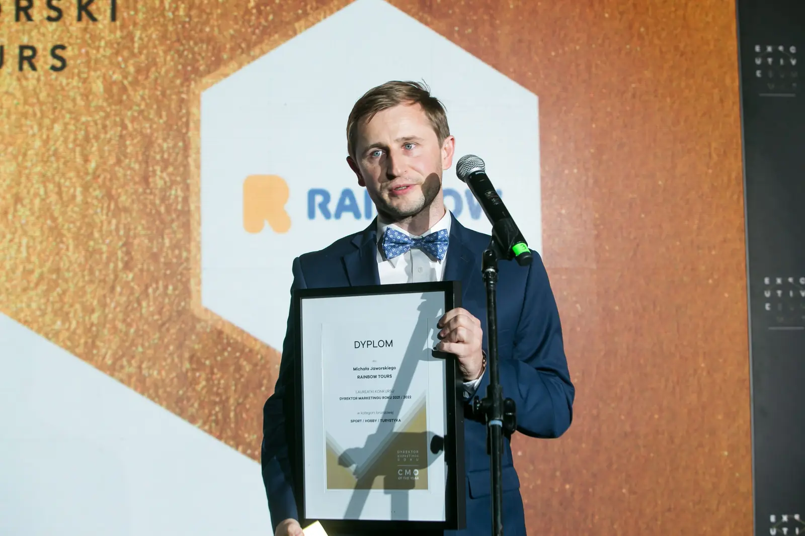 Uśmiechnięty laureat z muszką stoi przy mikrofonie z oprawionym dyplomem przed logo firmy — Gala DMR XI (2022)