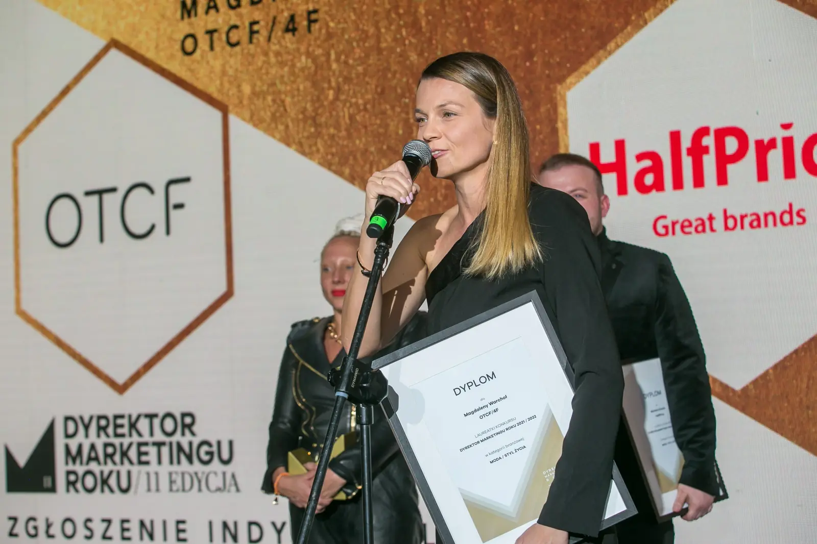 Laureatka z długimi włosami przemawia przy mikrofonie, trzymając oprawiony dyplom — Gala DMR XI (2022)