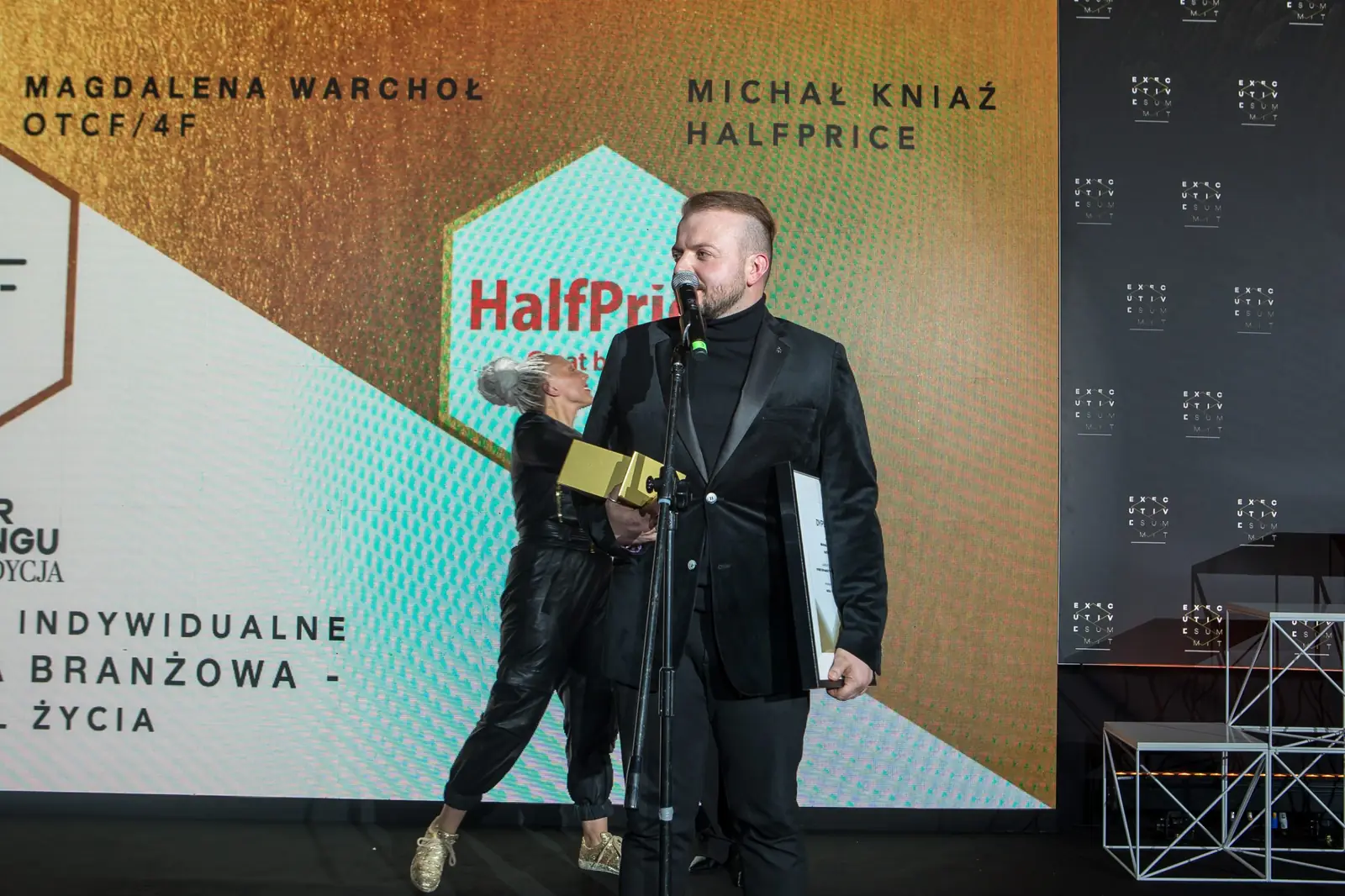Laureat w czarnym garniturze przemawia przy mikrofonie ze statuetką w kategorii moda — Gala DMR XI (2022)
