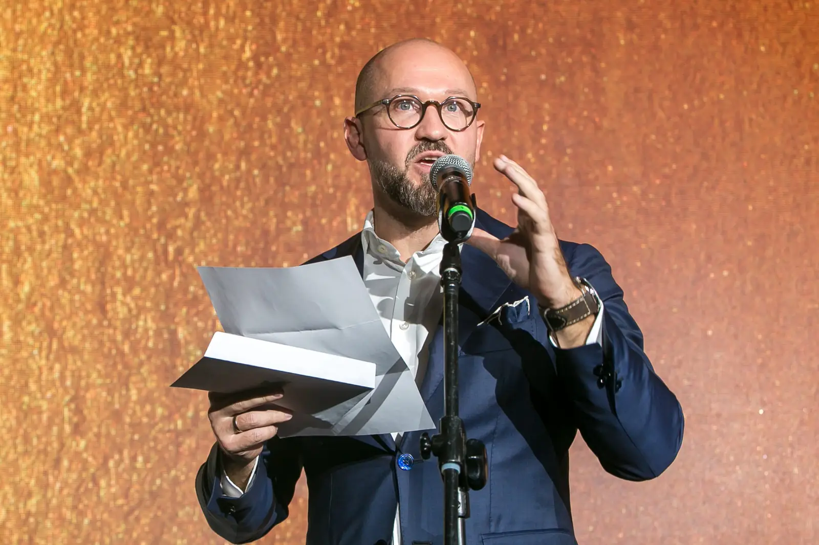 Mężczyzna w okrągłych okularach gestykuluje przy mikrofonie, trzymając otwartą kopertę — Gala DMR XI (2022)