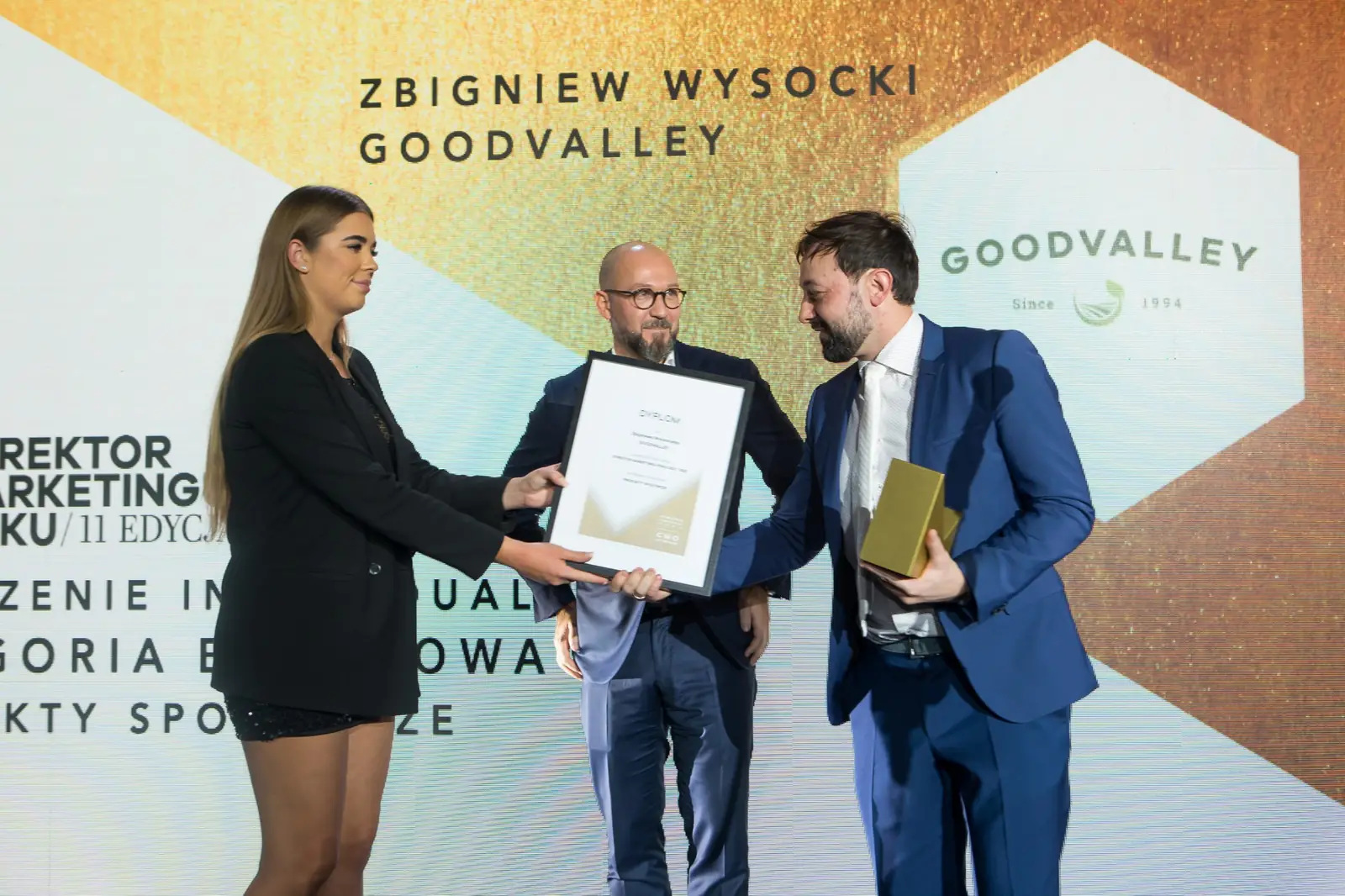 Asystentka w czarnej sukience podaje dyplom, a laureat w niebieskim garniturze trzyma statuetkę — Gala DMR XI (2022)