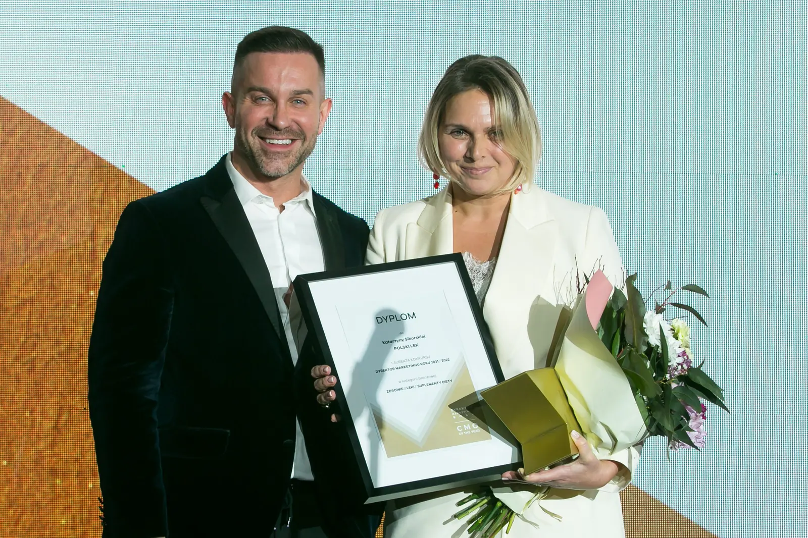 Laureatka w białym garniturze z dyplomem, kwiatami i statuetką pozuje z uśmiechniętym mężczyzną — Gala DMR XI (2022)