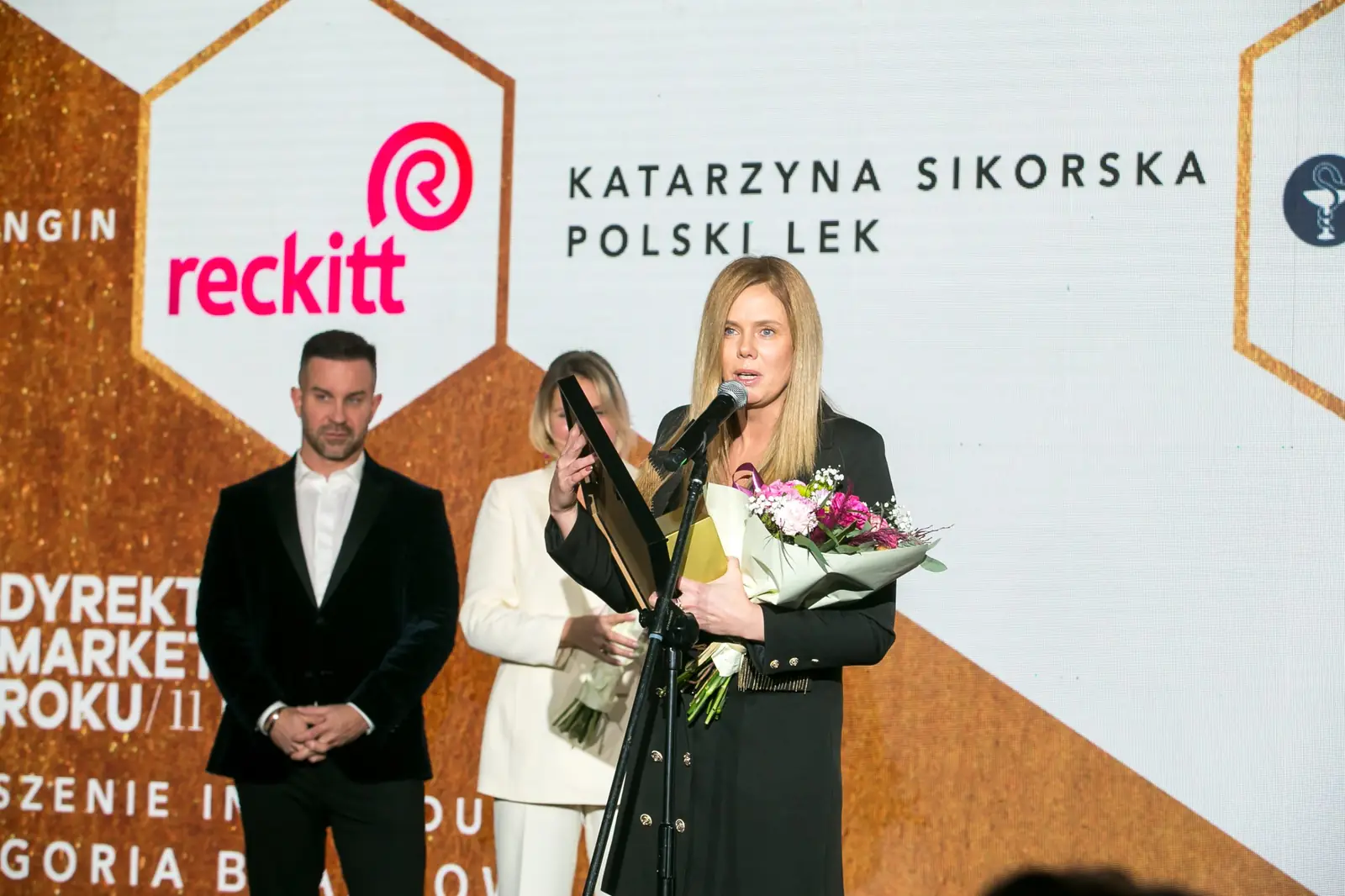 Laureatka z bukietem przemawia przy mikrofonie, za nią kobieta w białym garniturze i mężczyzna — Gala DMR XI (2022)