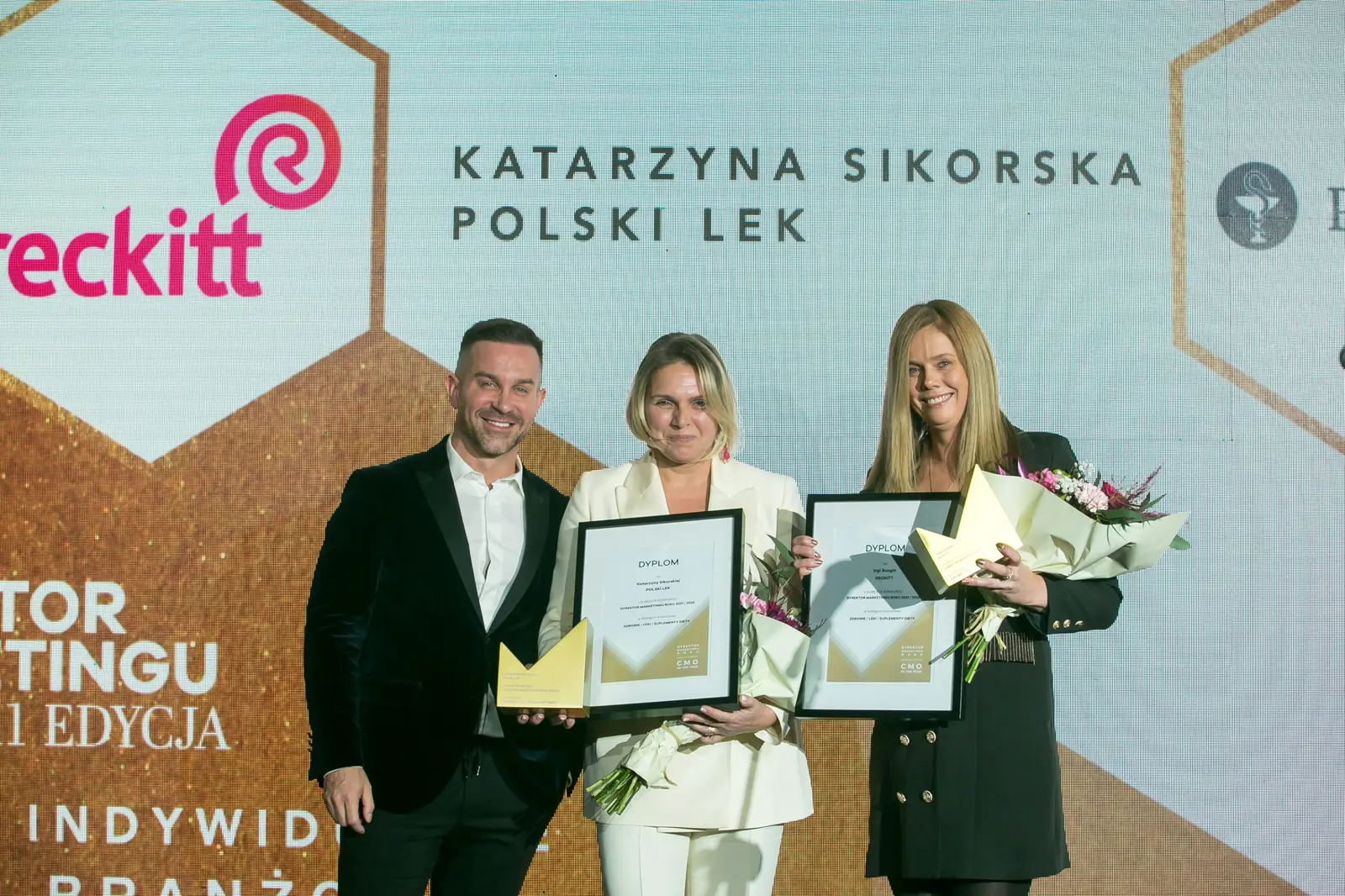Dwie laureatki z dyplomami, kwiatami i statuetkami pozują z mężczyzną w ciemnym garniturze — Gala DMR XI (2022)