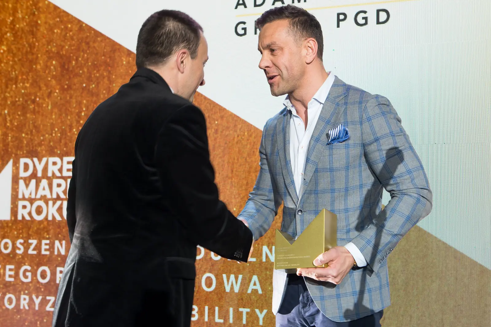 Wręczający w szarej marynarce w kratę ze statuetką w dłoni ściska rękę laureata — Gala DMR XI (2022)