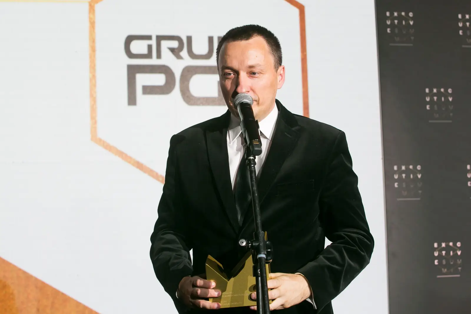 Laureat w czarnym garniturze i krawacie dziękuje, trzymając złotą statuetkę na wysokości pasa — Gala DMR XI (2022)