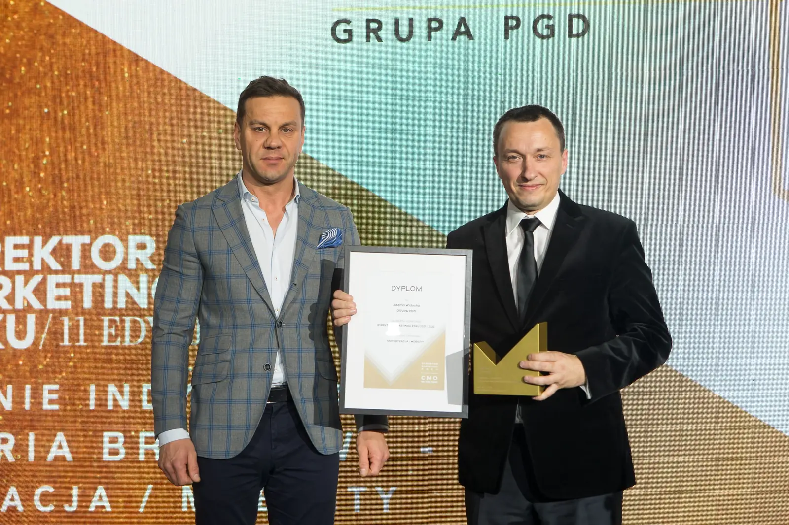 Dwóch mężczyzn w garniturach pozuje z oprawionym dyplomem i złotą statuetką przed ekranem — Gala DMR XI (2022)