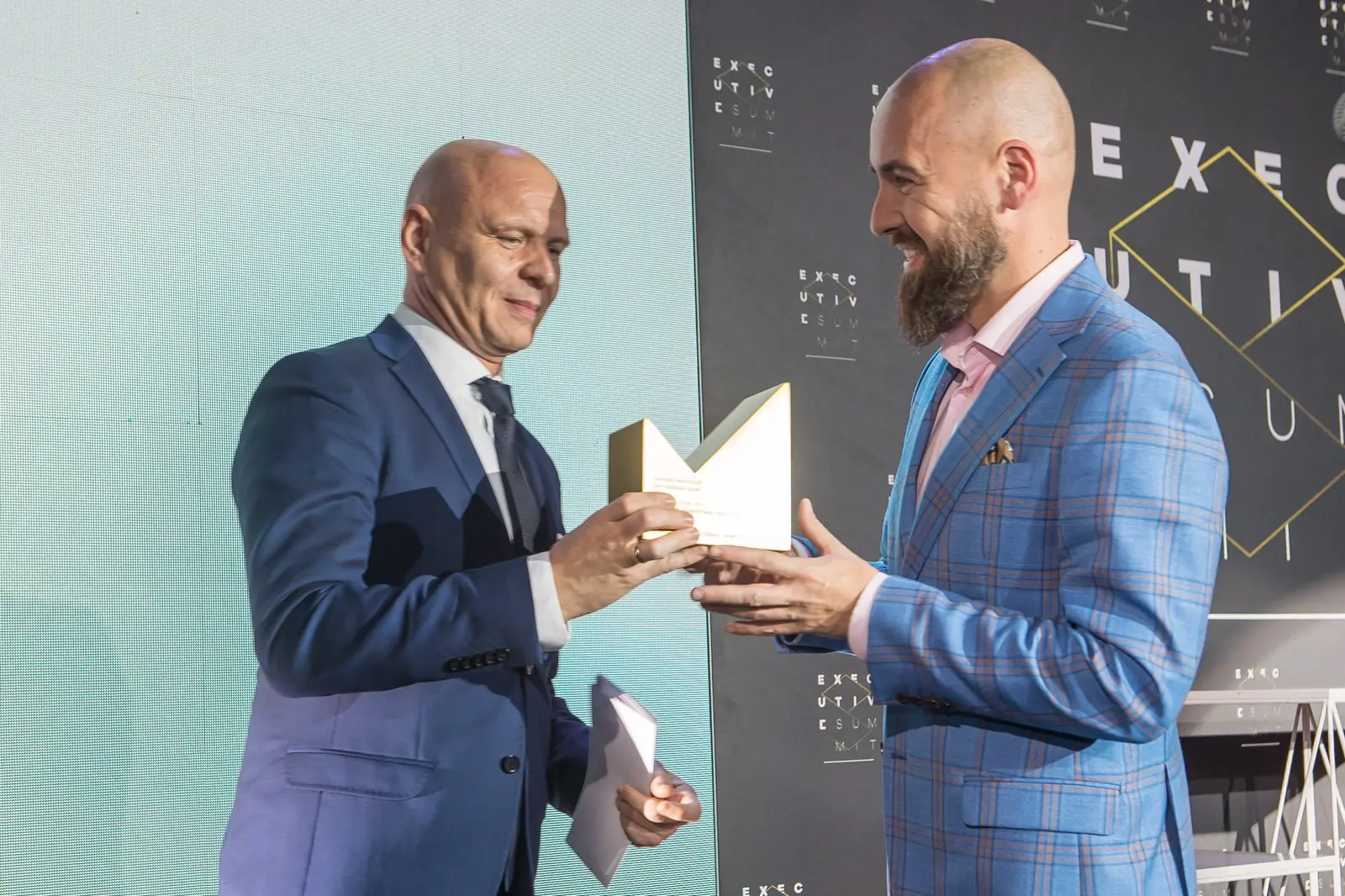 Wręczający w granatowym garniturze przekazuje statuetkę mężczyźnie w niebieskiej marynarce w kratę — Gala DMR XI (2022)