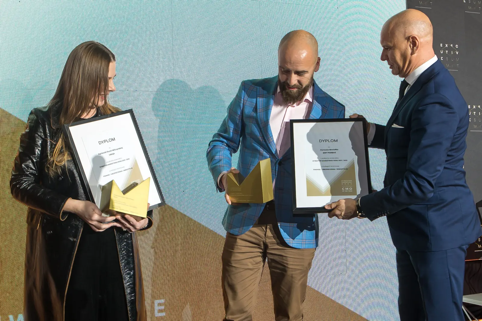 Wręczający podaje dyplom laureatowi w niebieskiej marynarce, obok laureatka z nagrodami — Gala DMR XI (2022)
