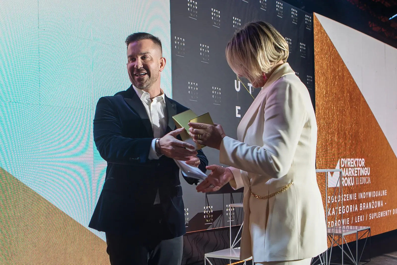 Uśmiechnięty mężczyzna w ciemnym garniturze wręcza statuetkę laureatce w białym garniturze — Gala DMR XI (2022)