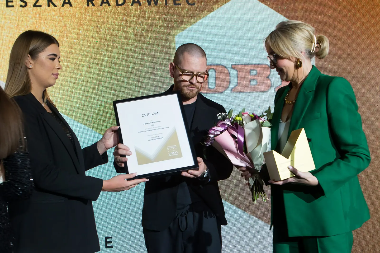 Asystentka wręcza dyplom mężczyźnie w czerni, obok laureatka w zieleni z kwiatami — Gala DMR XI (2022)