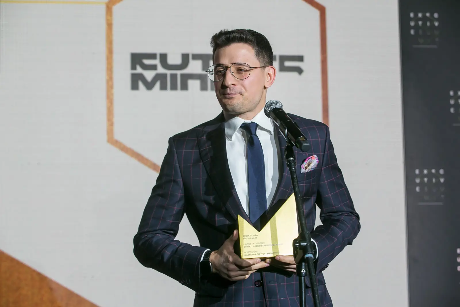Laureat w okularach i garniturze w kratę mówi do mikrofonu, trzymając złotą statuetkę — Gala DMR XI (2022)