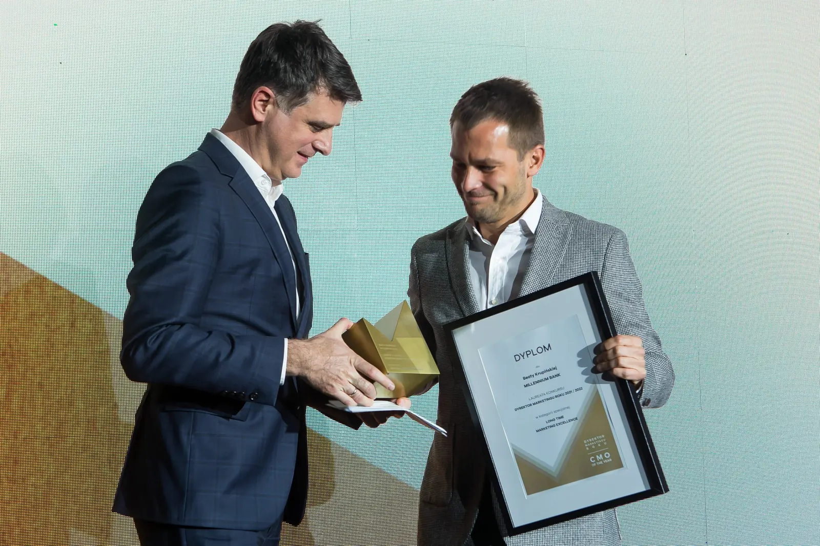 Laureat w szarym garniturze z dyplomem przyjmuje statuetkę, uśmiechając się szeroko — Gala DMR XI (2022)