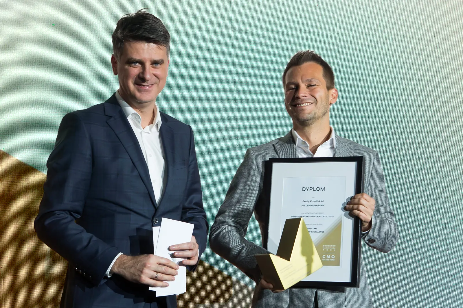 Laureat w szarym garniturze pozuje z dyplomem i statuetką obok mężczyzny z kopertą — Gala DMR XI (2022)