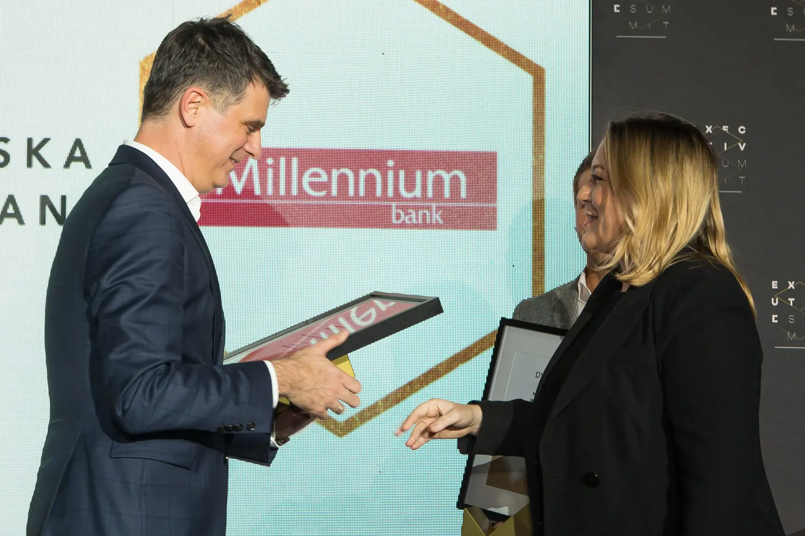 Mężczyzna w granatowym garniturze przekazuje oprawiony dyplom laureatce w czarnym żakiecie — Gala DMR XI (2022)