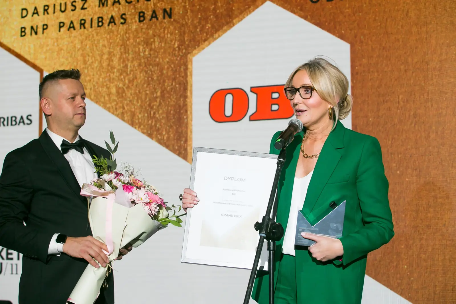 Laureatka w okularach trzyma dyplom i szklaną statuetkę przy mikrofonie, obok prowadzący — Gala DMR XI (2022)