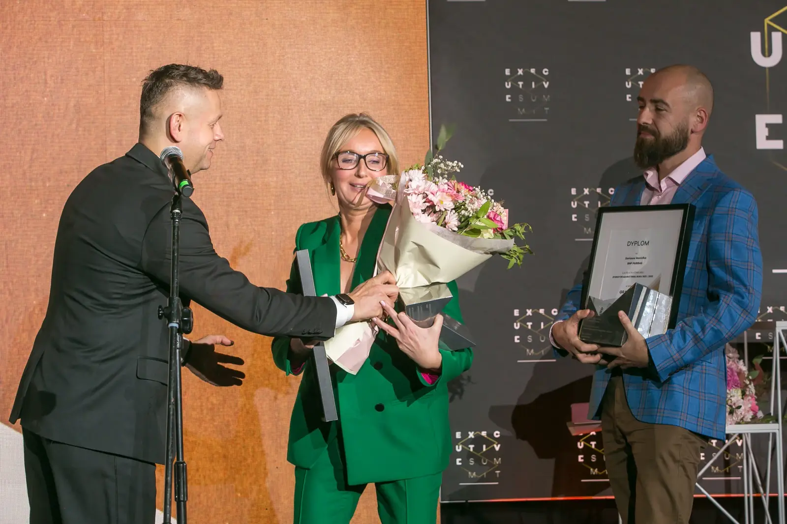 Prowadzący wręcza bukiet laureatce Grand Prix w zieleni, obok drugi laureat z dyplomem — Gala DMR XI (2022)