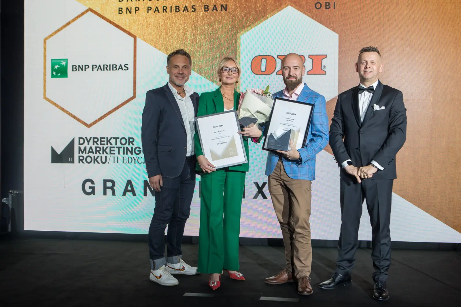 Szeroki kadr: laureaci Grand Prix i prowadzący stoją przed ekranem z napisem Grand Prix — Gala DMR XI (2022)