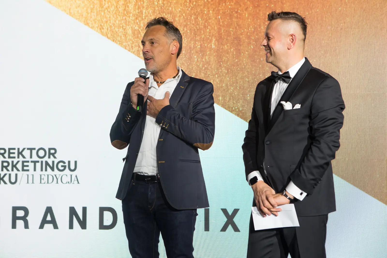 Współprowadzący z dłonią na sercu mówi do mikrofonu, a prowadzący uśmiecha się obok — Gala DMR XI (2022)