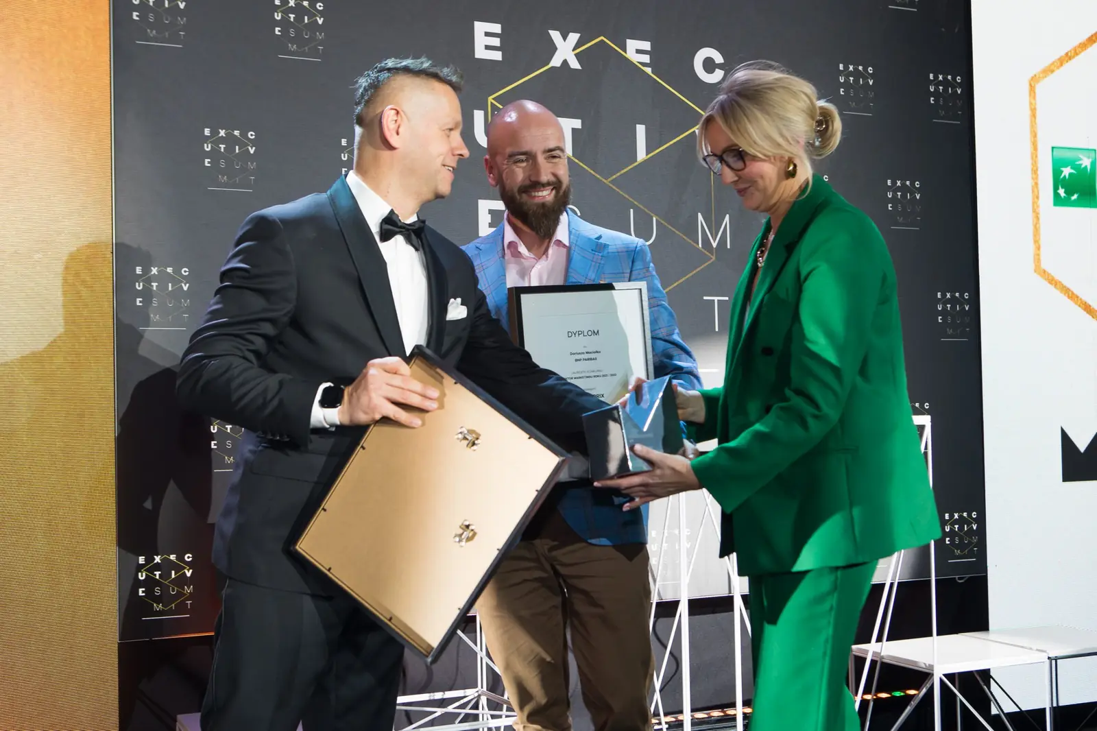 Laureat w niebieskiej marynarce z dyplomem i statuetką między prowadzącym a laureatką w zieleni — Gala DMR XI (2022)