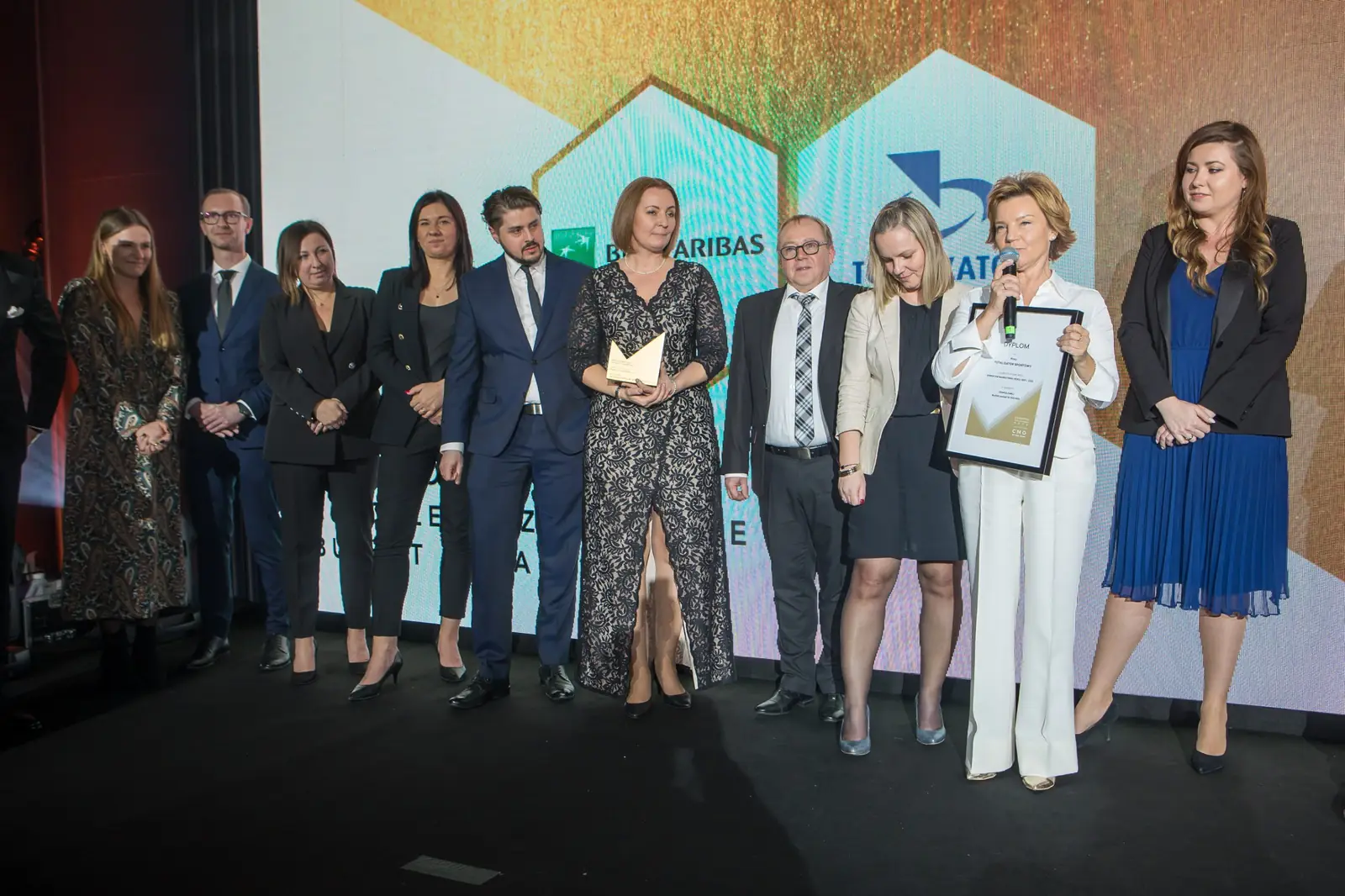 Szesnastoosobowa grupa laureatów w eleganckich strojach stoi w rzędzie na scenie — Gala DMR XI (2022)