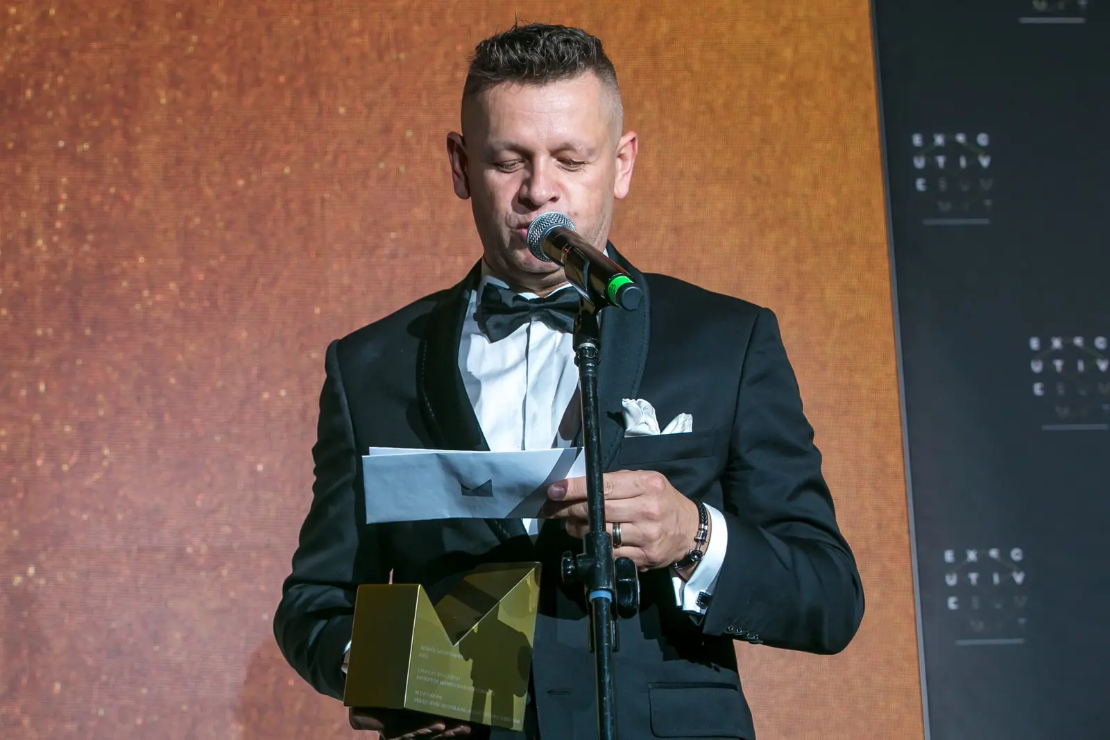 Prowadzący w smokingu czyta z kartki przy mikrofonie, trzymając złotą statuetkę — Gala DMR XI (2022)