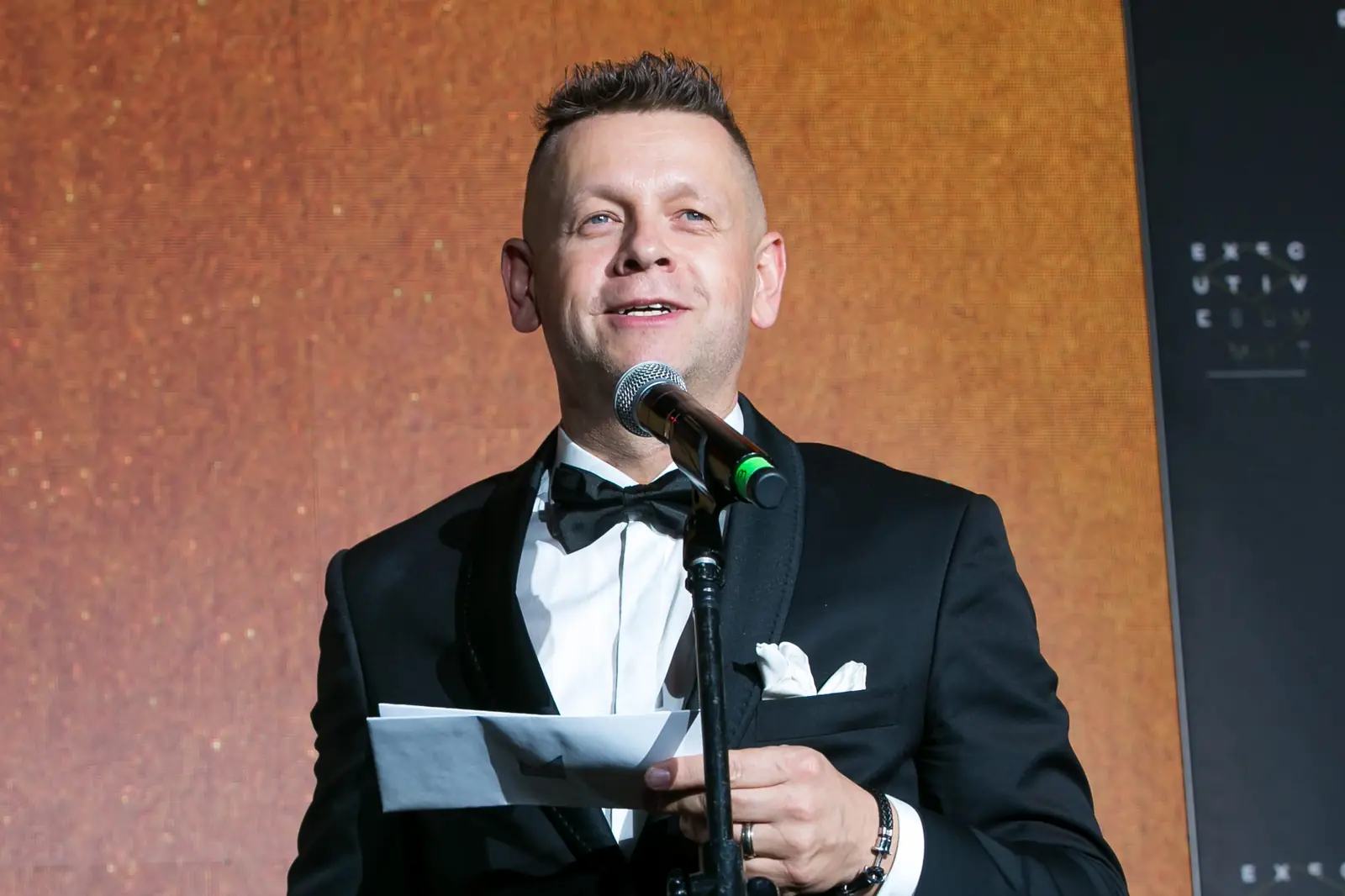Zbliżenie na uśmiechniętego prowadzącego z muszką, który zapowiada kolejnego laureata — Gala DMR XI (2022)