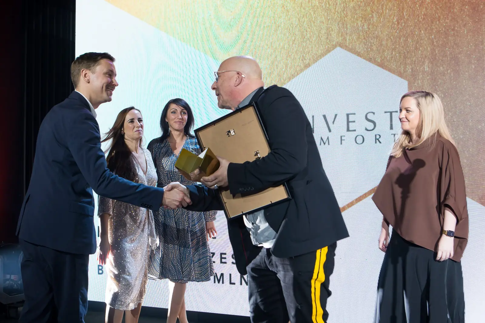 Laureat z dyplomem i statuetką ściska dłoń mężczyzny w granatowym garniturze, obok zespół — Gala DMR XI (2022)