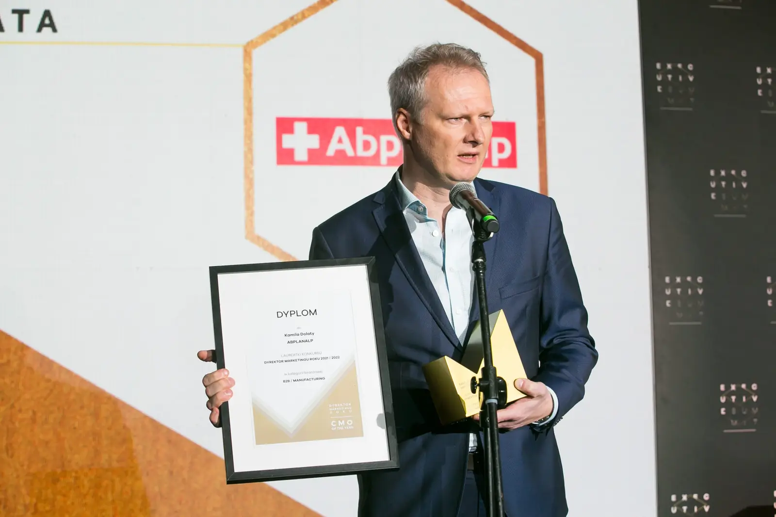 Laureat w granatowej marynarce i jasnej koszuli pokazuje dyplom przy mikrofonie — Gala DMR XI (2022)
