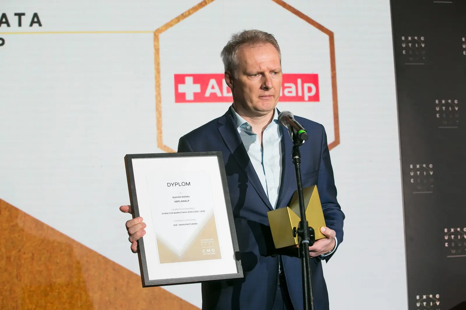 Laureat z oprawionym dyplomem i złotą statuetką przemawia przy mikrofonie na statywie — Gala DMR XI (2022)