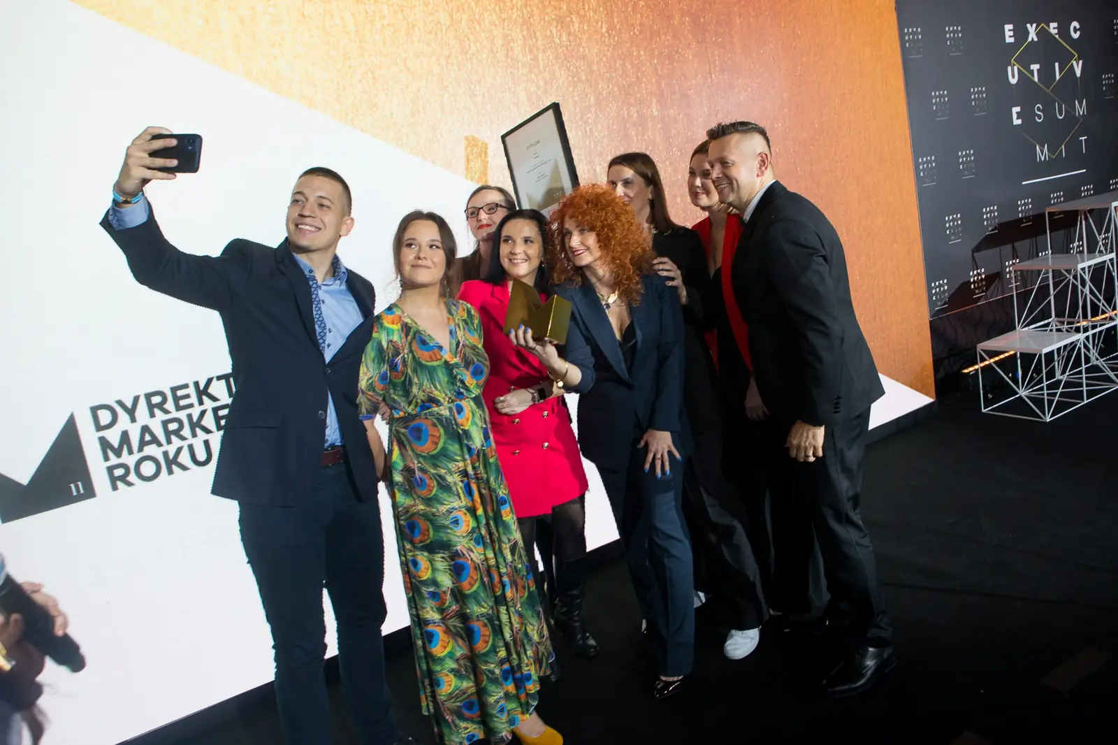 Mężczyzna w granatowym garniturze robi selfie z roześmianą grupą przy ściance konkursu — Gala DMR XI (2022)