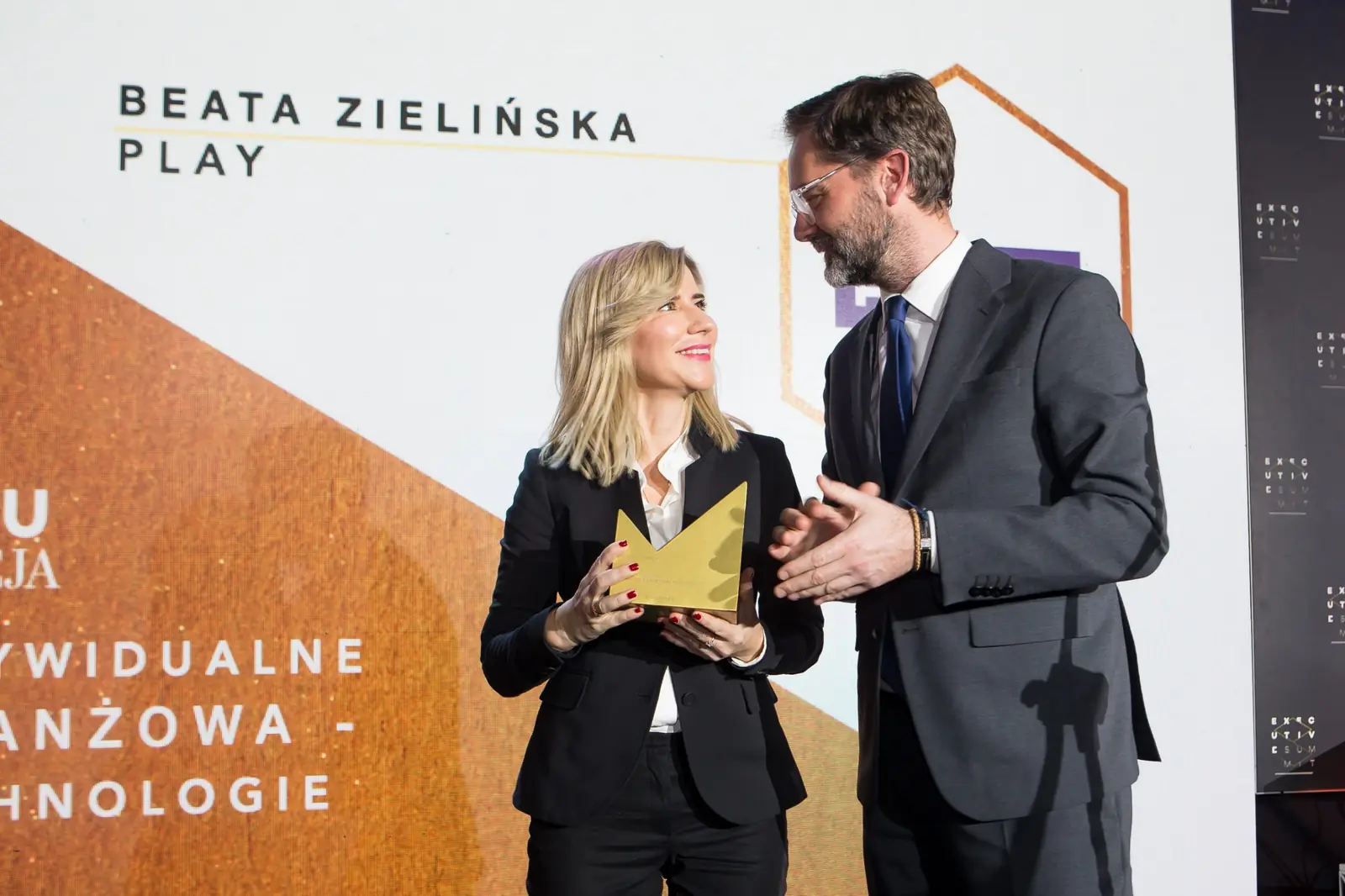 Wręczający bije brawo laureatce w czarnym żakiecie, która trzyma złotą statuetkę — Gala DMR XI (2022)