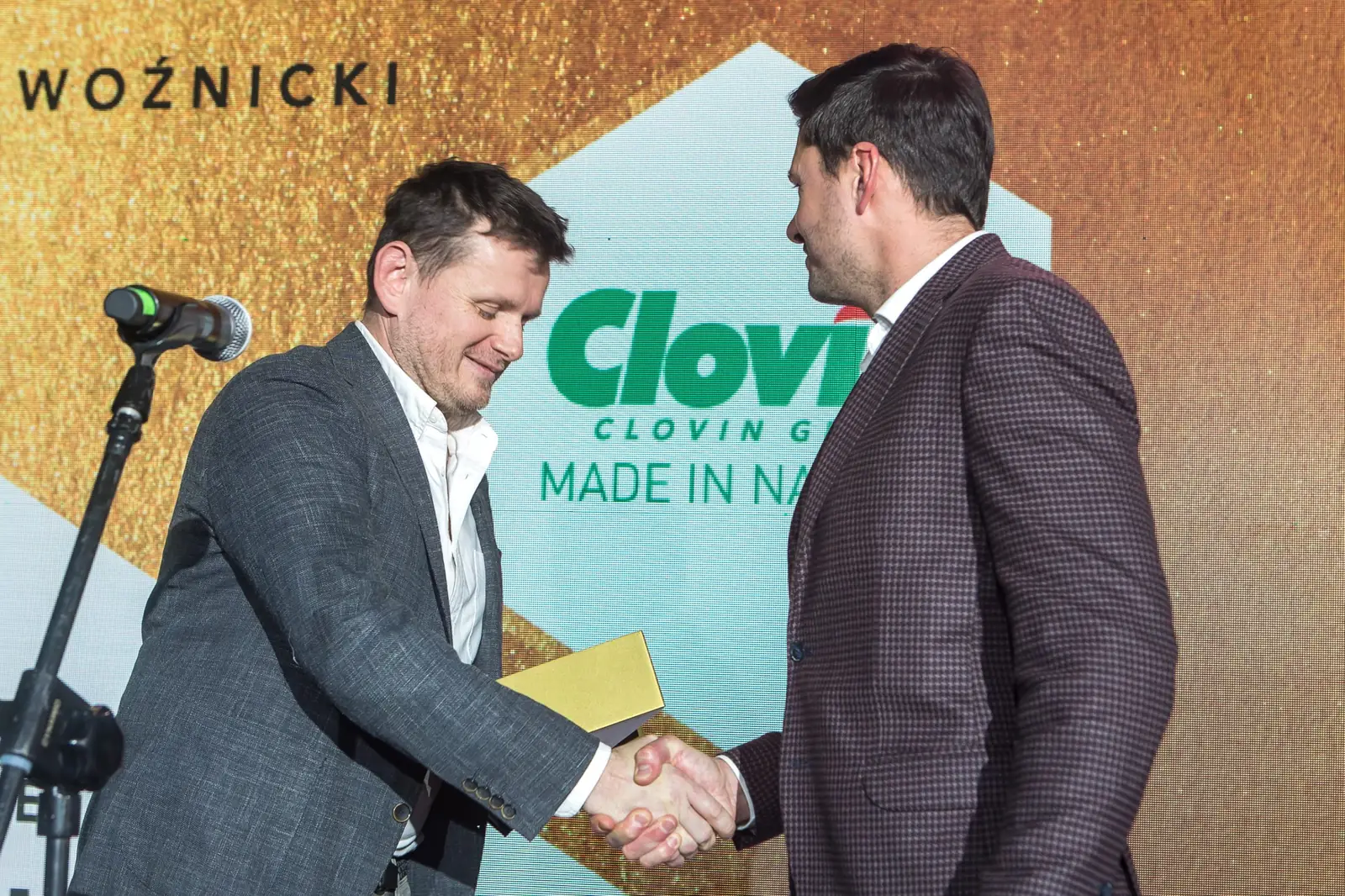Laureat w bordowej marynarce w kratę ściska dłoń wręczającego stojącego przy mikrofonie — Gala DMR XI (2022)