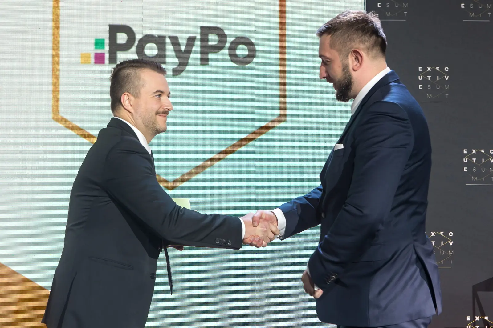 Mężczyzna z wąsem ściska dłoń laureata w granatowym garniturze przed logo firmy — Gala DMR XI (2022)