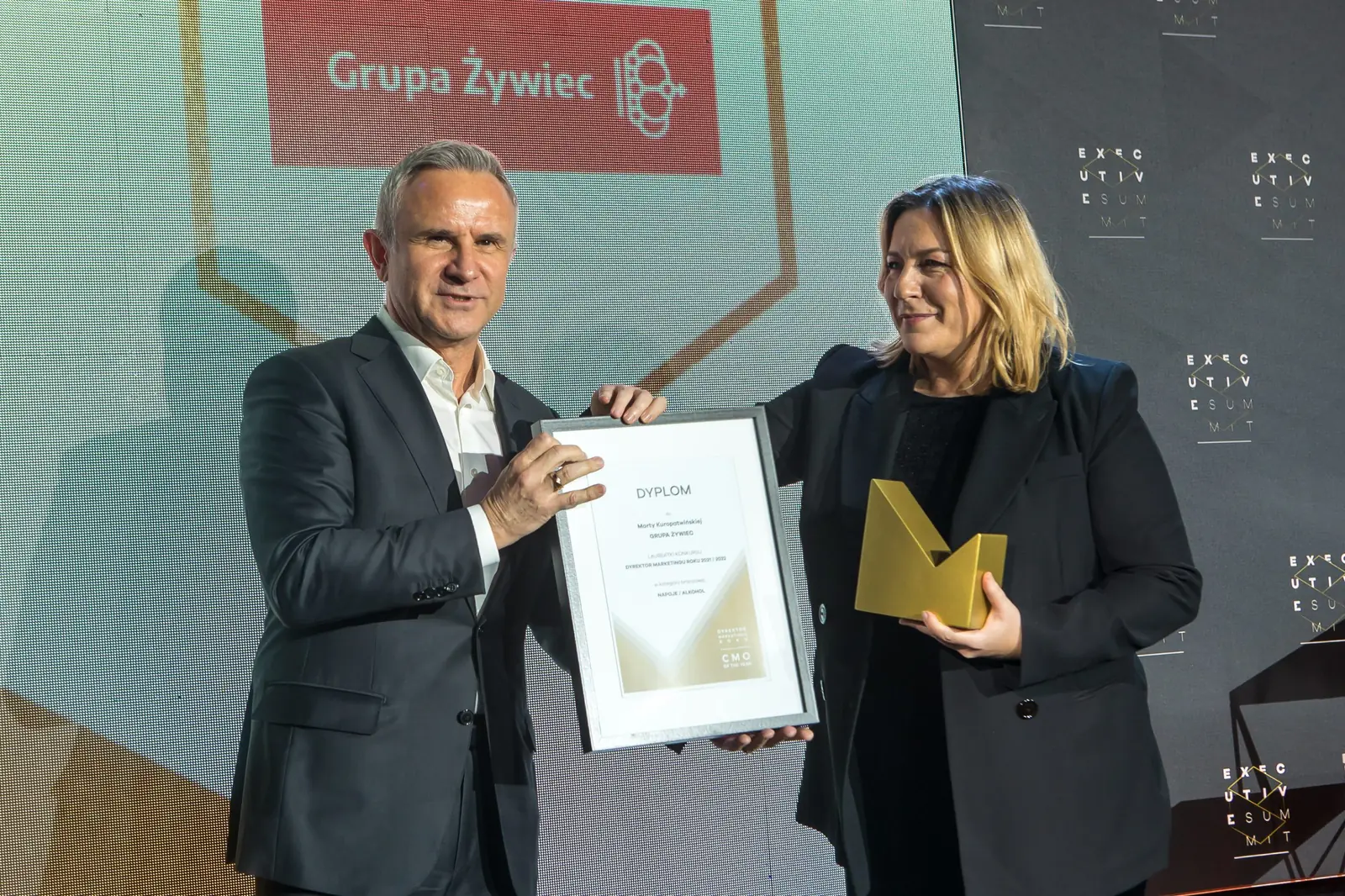 Szpakowaty wręczający trzyma dyplom, a laureatka w czarnej marynarce ściska złotą statuetkę — Gala DMR XI (2022)
