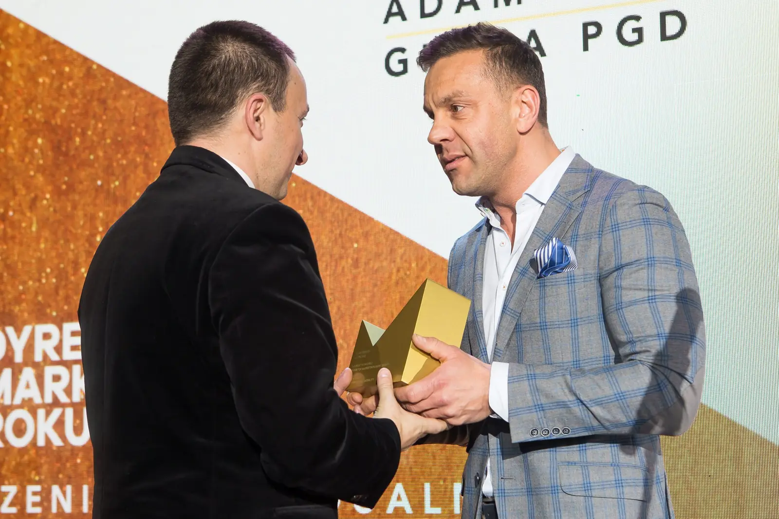 Mężczyzna w marynarce w kratę z poszetką wręcza statuetkę laureatowi w czarnym garniturze — Gala DMR XI (2022)