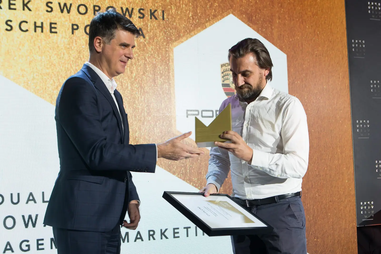Wręczający wyciąga dłoń do brodatego laureata, który trzyma już statuetkę i dyplom — Gala DMR XI (2022)