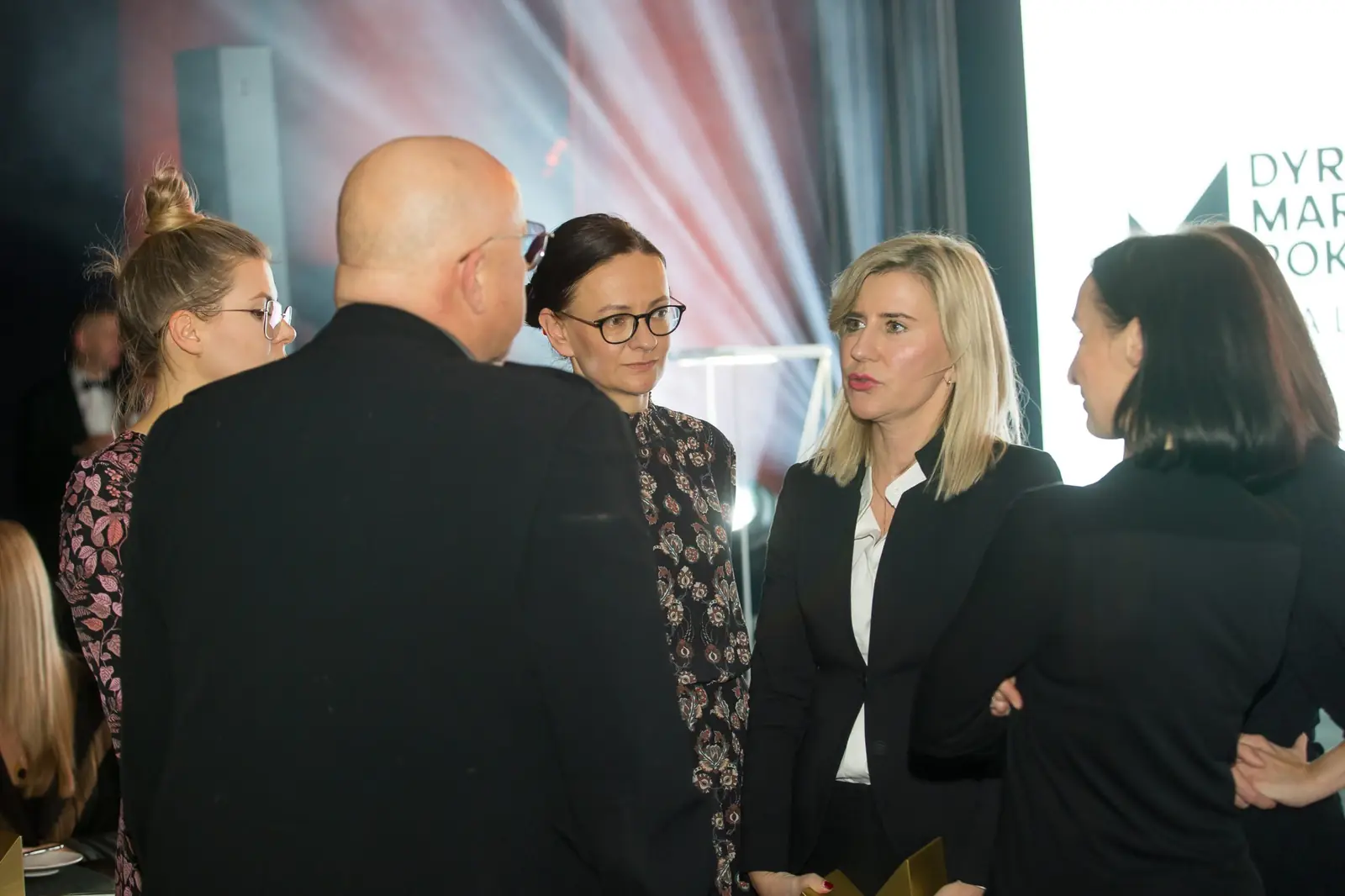 Goście rozmawiają w małych grupkach przy scenie rozświetlonej białymi reflektorami — Gala DMR XI (2022)