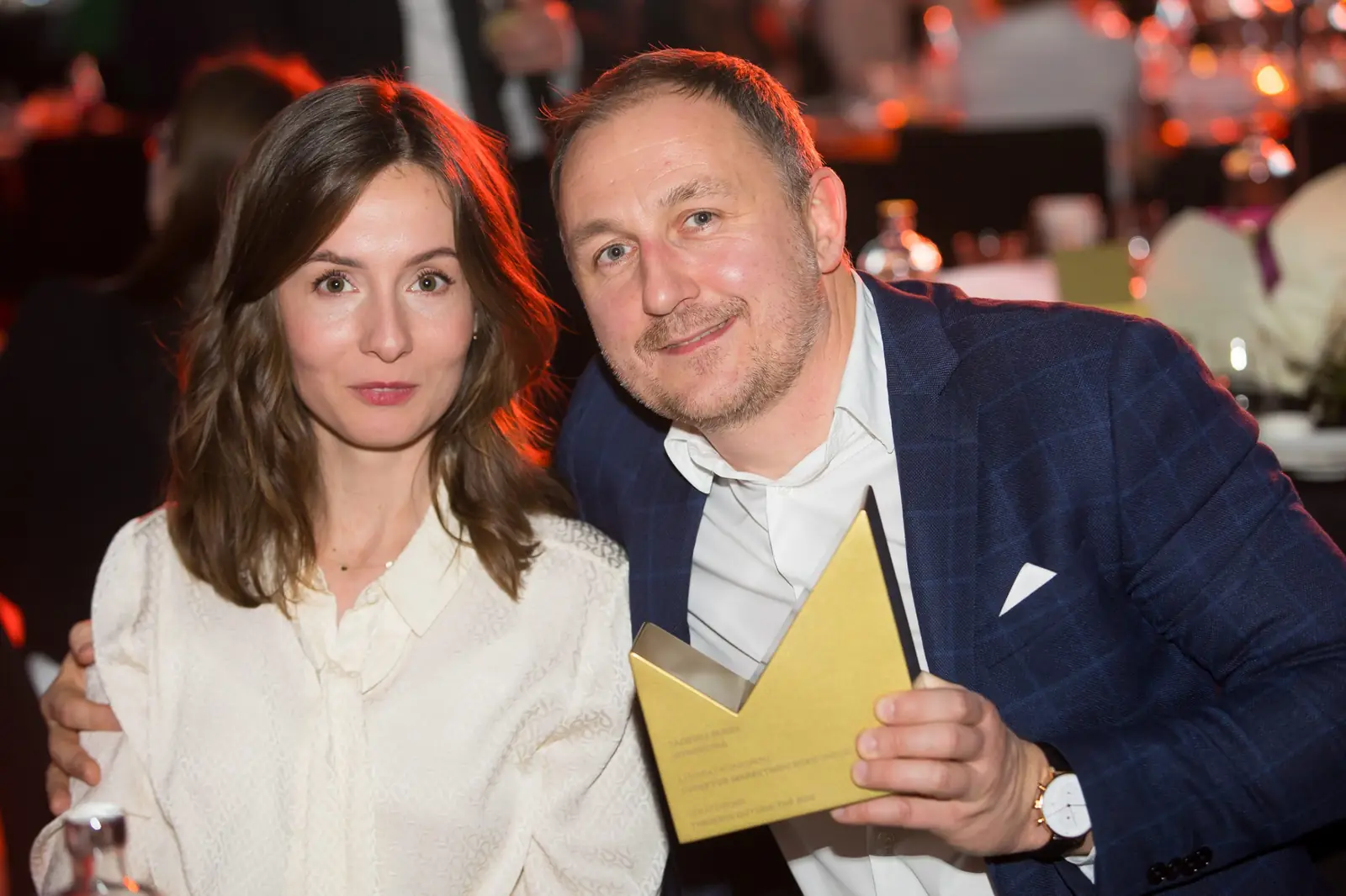 Mężczyzna w granatowym garniturze ze statuetką i kobieta w białej bluzce pozują przy stolikach — Gala DMR XI (2022)