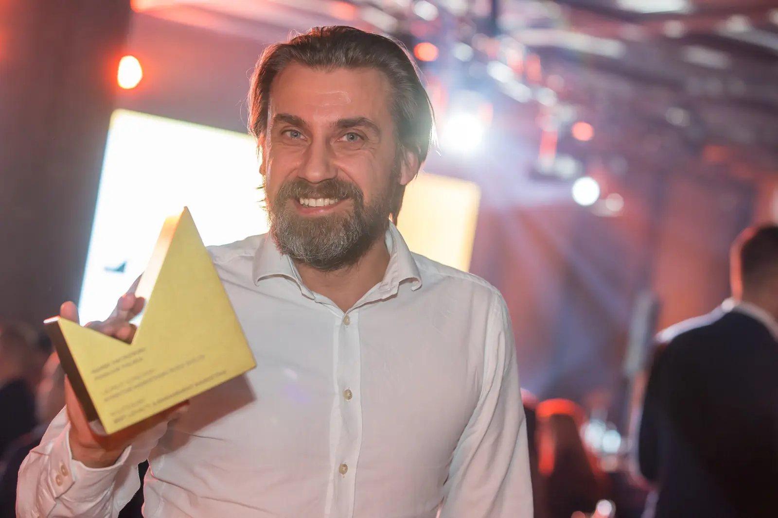 Brodaty laureat w białej koszuli z uśmiechem prezentuje złotą statuetkę w sali — Gala DMR XI (2022)