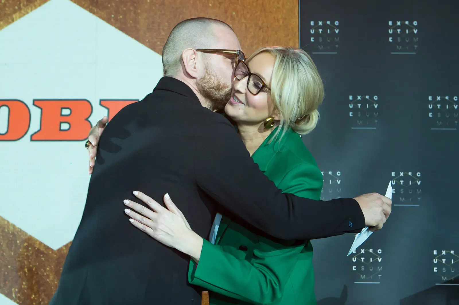 Mężczyzna w czerni serdecznie ściska laureatkę w zielonym żakiecie przed logo firmy — Gala DMR XI (2022)