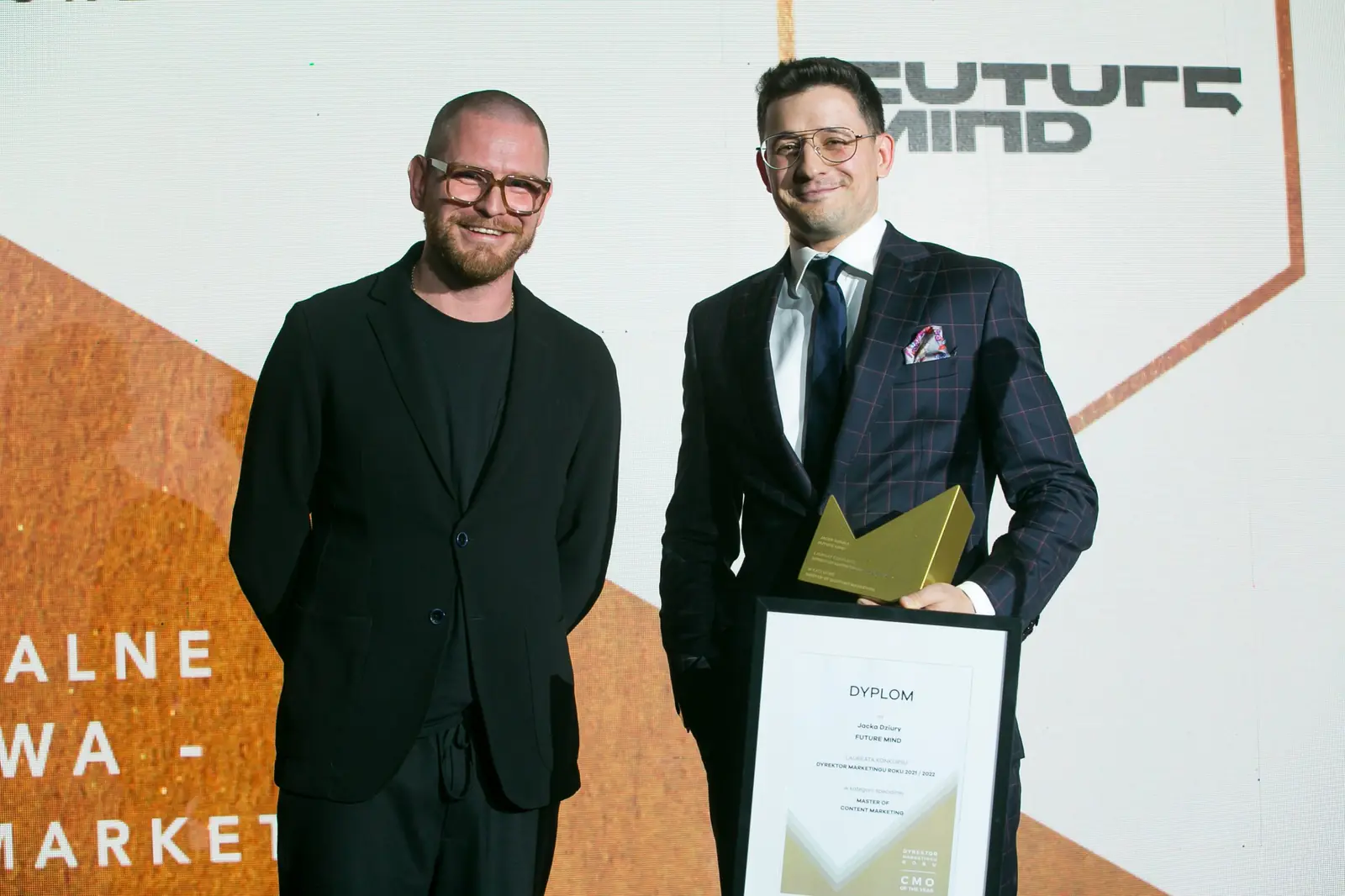 Laureat z dyplomem i statuetką pozuje obok wręczającego w czarnym stroju i okularach — Gala DMR XI (2022)