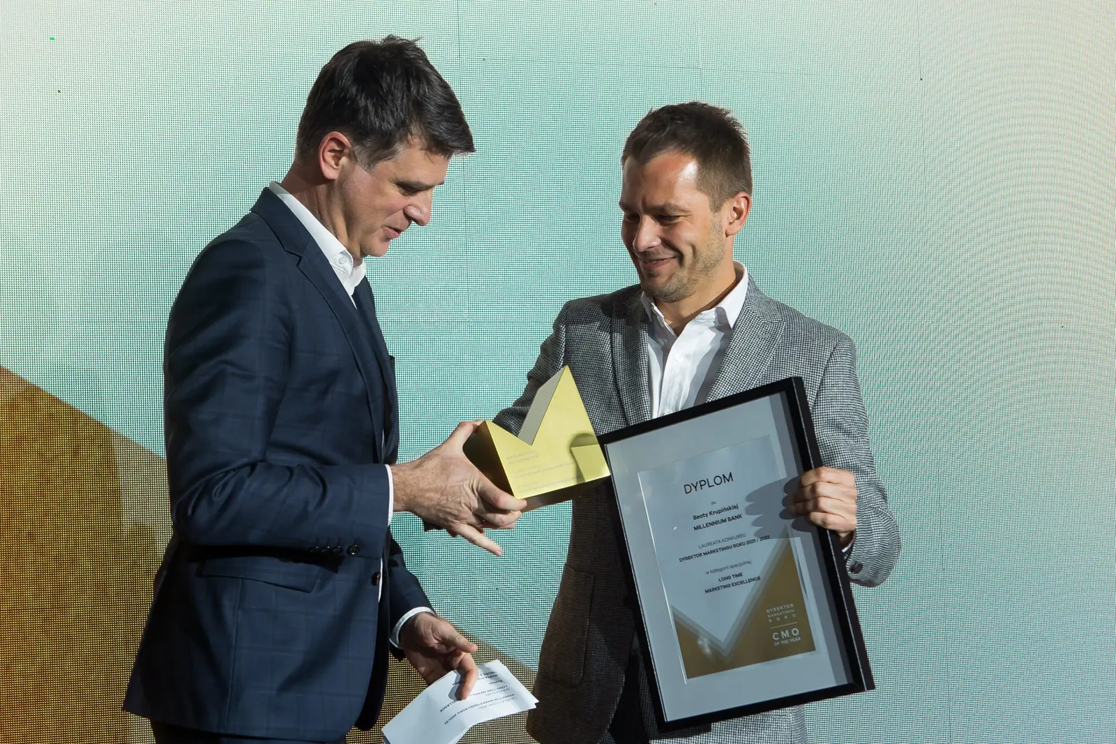 Mężczyzna w granatowym garniturze z kopertą wręcza statuetkę laureatowi w szarym garniturze — Gala DMR XI (2022)