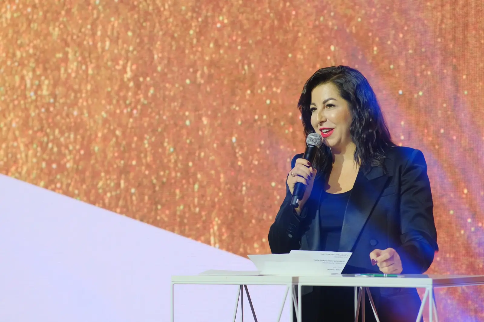 Kobieta w ciemnej marynarce przemawia do mikrofonu przy mównicy na tle złocistej ściany — Gala DMR XII (2023)
