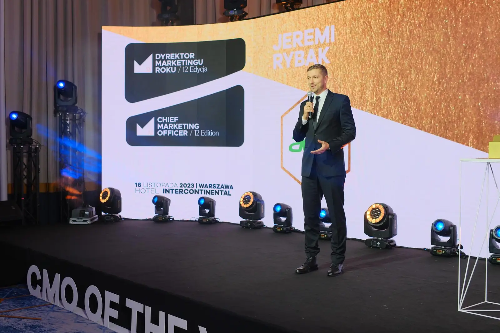 Laureat przemawia z mikrofonem na szerokiej scenie z logo konkursu i rzędem reflektorów — Gala DMR XII (2023)