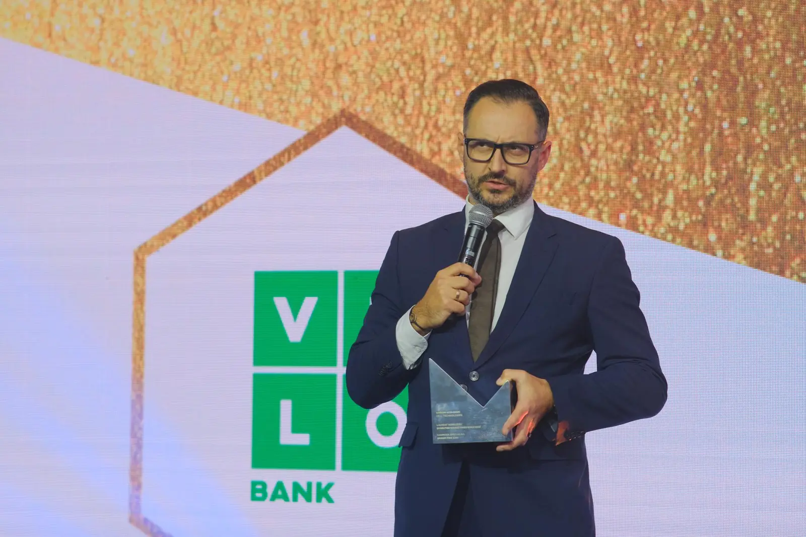 Laureat w okularach dziękuje do mikrofonu, trzymając statuetkę przed ekranem z zielonym logo — Gala DMR XII (2023)