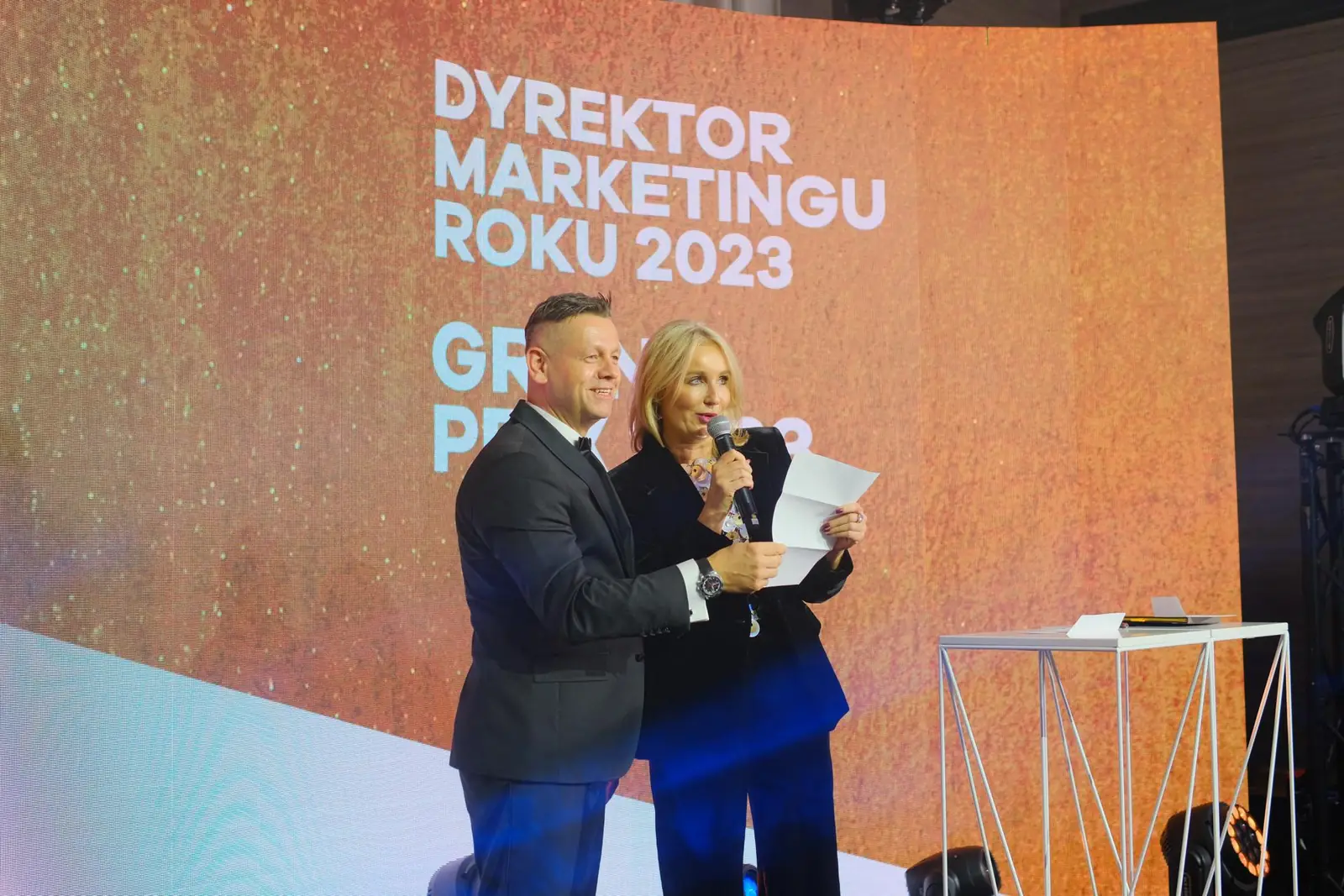 Prowadząca odczytuje zwycięzcę Grand Prix, a uśmiechnięty prowadzący stoi tuż obok — Gala DMR XII (2023)