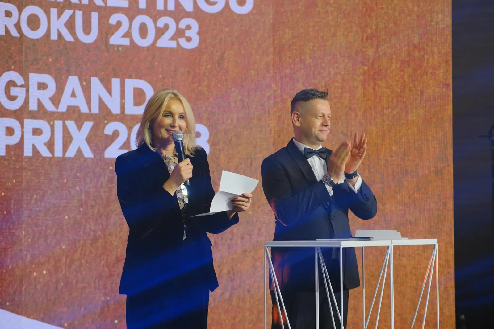 Prowadząca w granatowym kostiumie mówi do mikrofonu, a prowadzący bije brawo przy mównicy — Gala DMR XII (2023)
