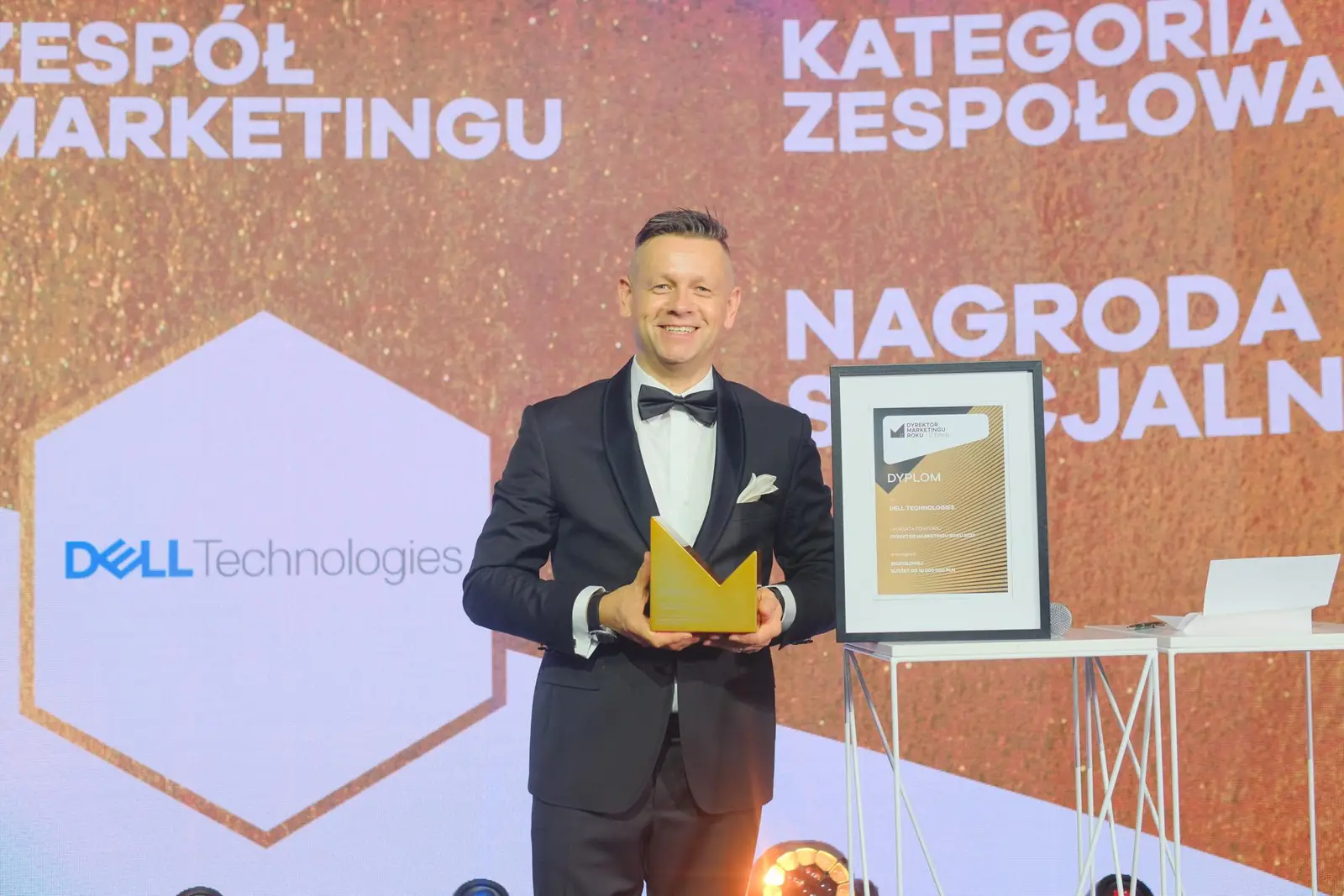 Prowadzący w smokingu z uśmiechem prezentuje złotą statuetkę i dyplom Nagrody Specjalnej — Gala DMR XII (2023)